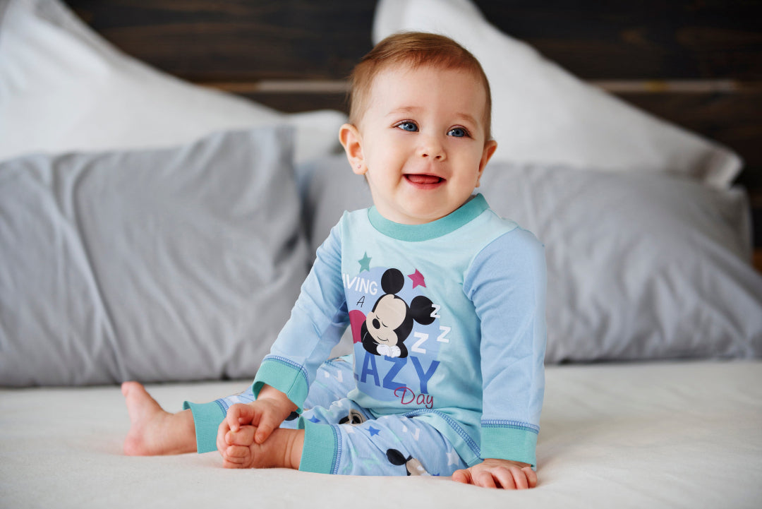 Baby wearing pajamas with a cartoon character, sitting on a bed.